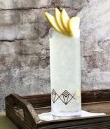 Cocktail in a tall glass with lemon slices on a wooden tray against a stone wall.