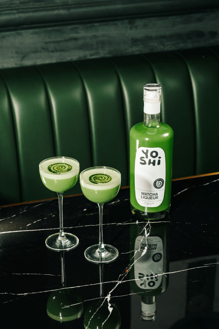Green bottle labeled 'Yoshi' with two matching cocktails on a reflective surface.
