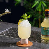A pale yellow cocktail filled with crushed ice in a clear coupe-style glass, garnished with mint leaves and a lemon wedge, sitting on a coaster on a bar counter. A bottle of Liber & Co. Syrup is blurred in the background.
