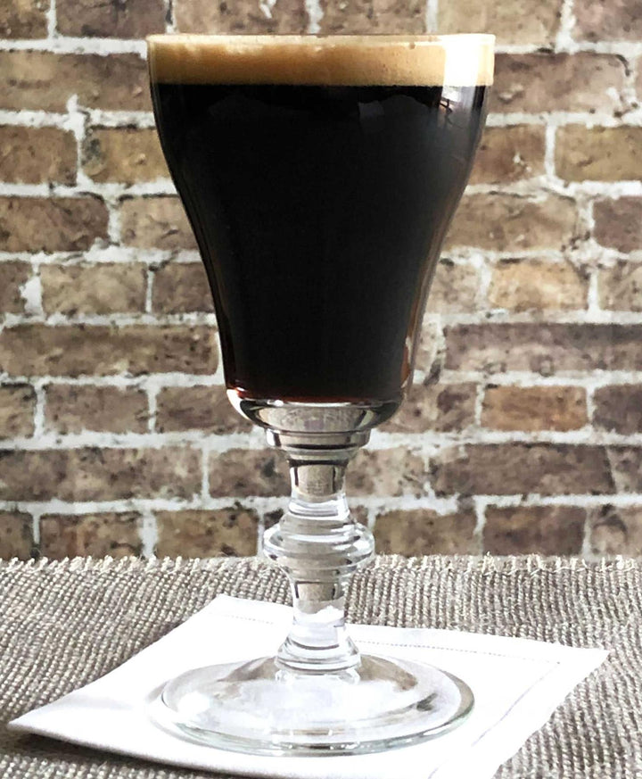 Glass of dark liquid with a foamy top on a textured surface with a brick wall background