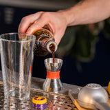 Bartender pouring dark liquid from a bottle into a shaker on a bar counter.