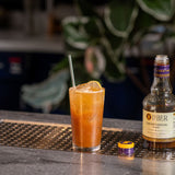 Cocktail with a lemon twist and bottle of Liber & Co Caramelized Fig Syrup on a bar counter.