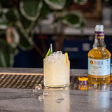 Cocktail with ice and lemon wedge next to a bottle of Liber Classic Gum Syrup on a bar.