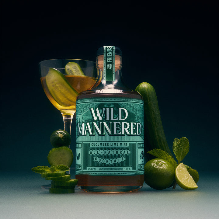 A bottle of Wild Mannered cucumber lime mint cocktail spirit centered on a dark studio background, surrounded by a filled cocktail glass, limes, mint leaves, and a cucumber.