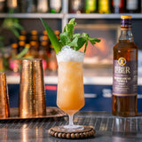 Cocktail with ice and garnish next to a bottle of Liber & Co. Syrup on a bar.