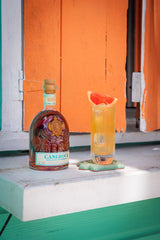 Canerock bottle and cocktail with grapefruit garnish on a wooden surface with an orange wall in the background.