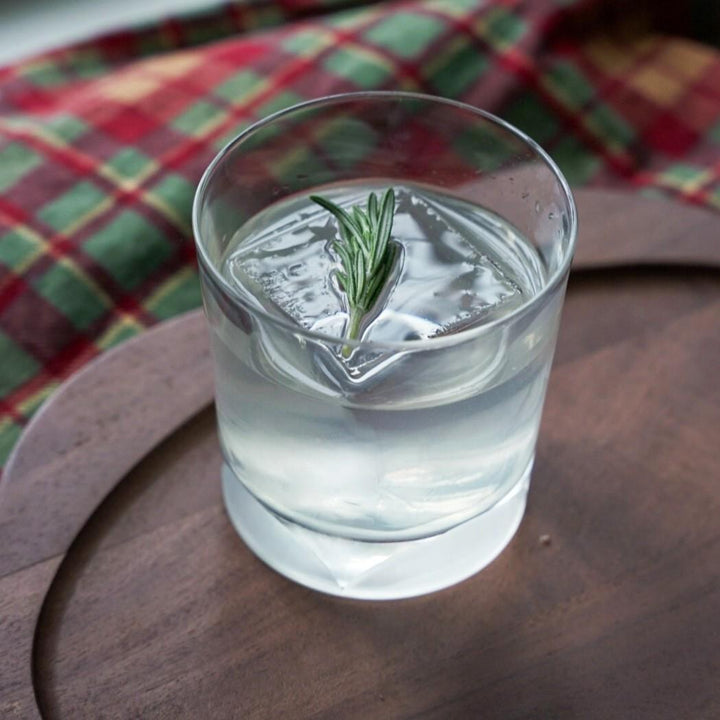 Clear glass of water with a sprig of rosemary on a wooden surface with a plaid cloth in the background.