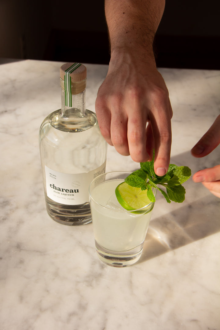 Person garnishing a cocktail with mint leaves next to a bottle of Chareau on a marble surface.