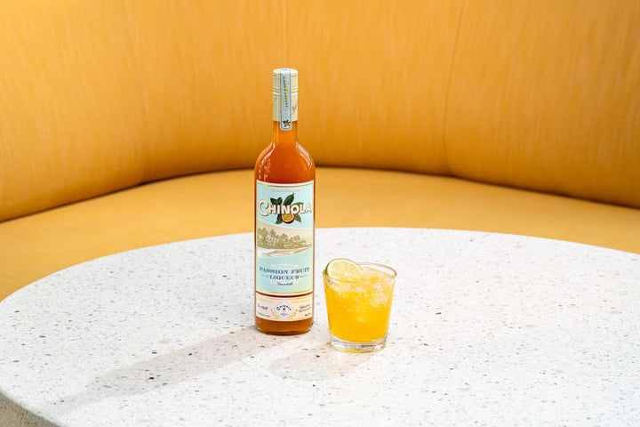 Bottle of Chinola with a glass of orange juice on a textured surface with a yellow background