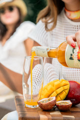 Person pouring a drink from a bottle into a glass with mangoes and passion fruits on a wooden board.