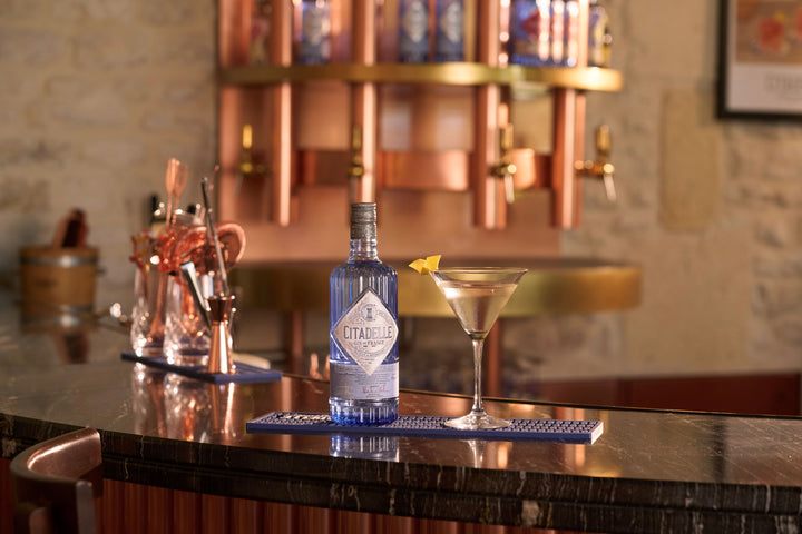 Bottle of Citadel gin with a cocktail on a bar counter, featuring a blurred bar background.