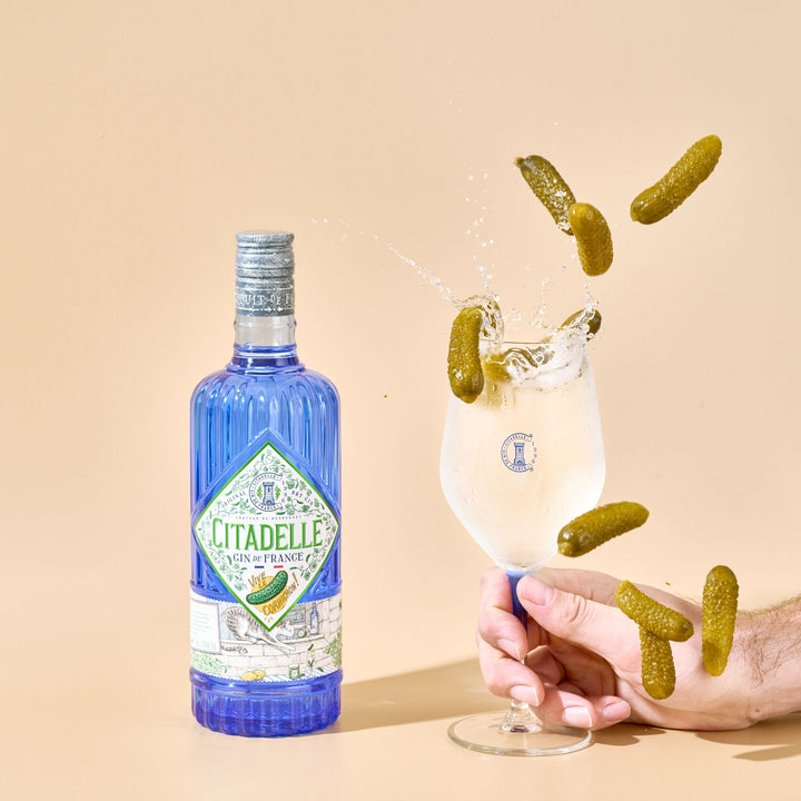 Citadelle gin bottle with a glass of gin and tonic, pickles, and a hand on a beige background