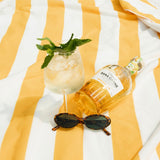 Bottle of Bana Canne rum, a glass with a drink, and sunglasses on a yellow and white striped fabric.