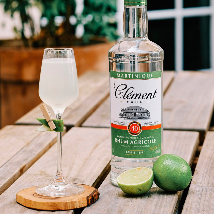 Bottle of Clément Rhum Agricole with a glass of white drink and limes on a wooden surface.