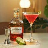 Bottle of Créole Shrubb with a cocktail glass filled with a red drink, lime wedges, and a jigger on a table.