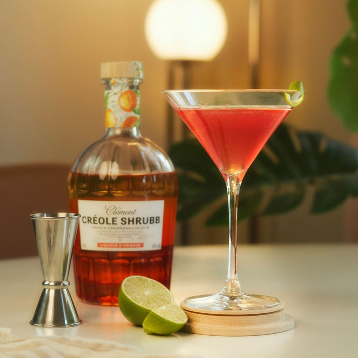 Bottle of Créole Shrubb with a cocktail glass filled with a red drink, lime wedges, and a jigger on a table.