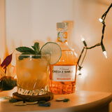 Glass of orange cocktail with lime and mint garnish next to a bottle of alcohol on a table with string lights in the background.