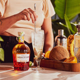 Person making a cocktail with Creole Shrubs bottle and ingredients on a table.