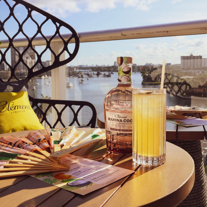 Bottle of Mahina Coc and a glass of yellow drink on a table with a scenic background