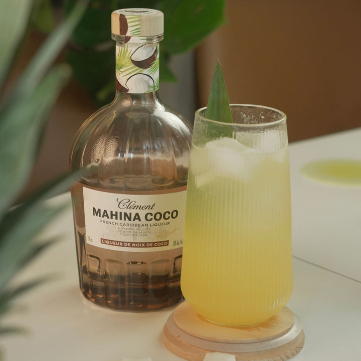 Bottle of Clément Mahina Coco liqueur with a glass of a yellow cocktail on a table.