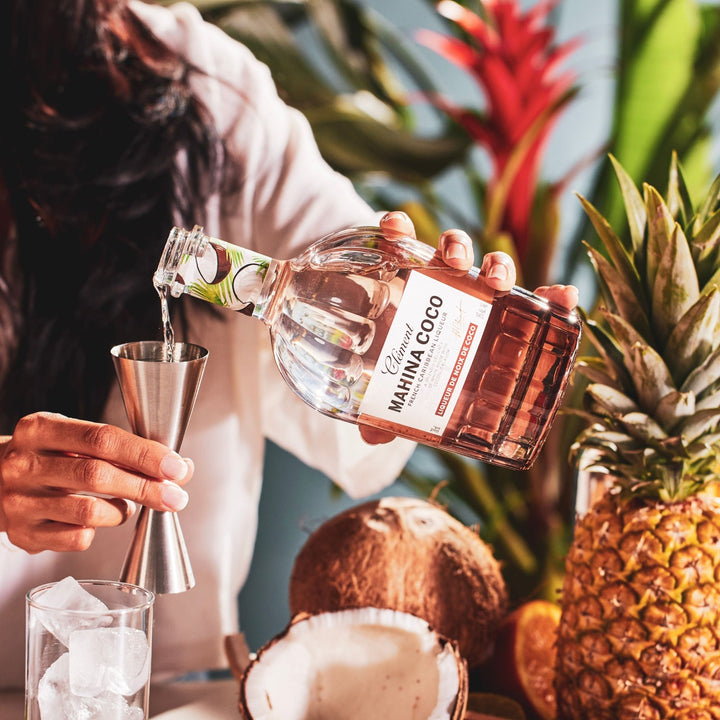 Person pouring a clear liquid from a bottle labeled 'Mahina Coco' into a cocktail shaker with tropical fruits and plants in the background.