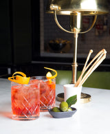 Two red cocktails with garnishes on a table with a brass lamp in the background.