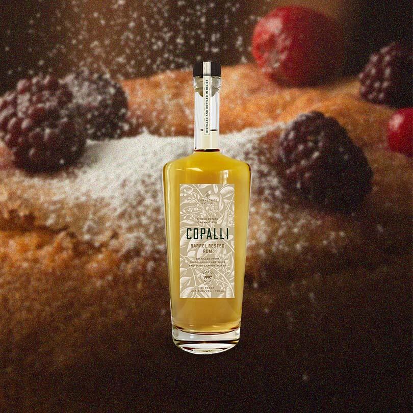 An elegant looking bottle of caramel colored liquid with a label that reads Copalli Barrel Rested Rum against a backdrop of illustrated rainforest flora and fauna.  All set against an image of a delicious cake with berries on top, evoking the flavor notes of blackberry, vanilla, spiceed cinnamon and nutmeg.