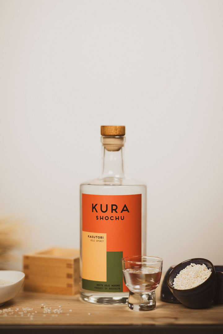 Kura Shochu bottle on a wooden surface with a glass of liquid and small bowls.