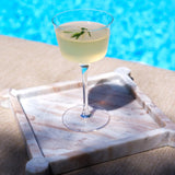 Cocktail in a glass with a sprig of rosemary on a marble tray by a pool.