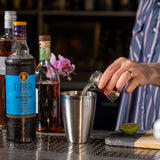 Person pouring a drink into a cocktail shaker with bottles and a lime in the background