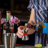Person pouring a drink into a cocktail shaker with bottles and a lime in the background