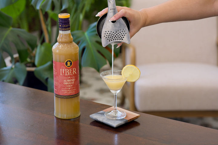 Person pouring a drink from a bottle labeled 'Liber' into a glass with a lemon slice, with a plant and chair in the background.