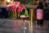 Cocktail with a garnish on a bar counter next to a bottle of Liber & Co Real Grenadine 