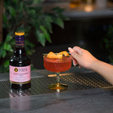 Bottle of Liber & Co Pacific Strawberry Syrup next to a cocktail with a hand holding it on a bar.