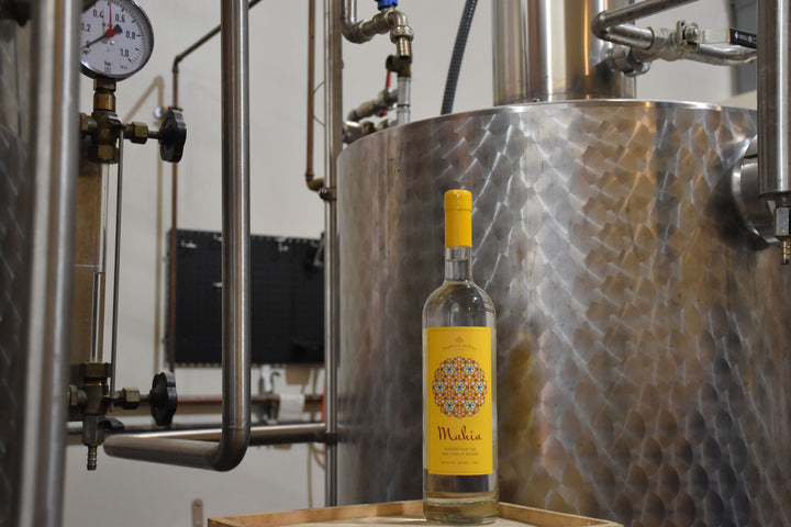 Bottle of Mahia Fig Liqueur wine with a yellow label in front of a large metallic tank in a winery setting.