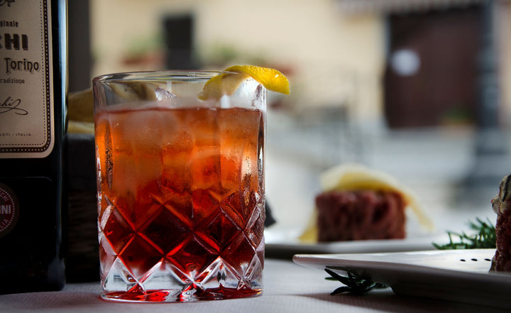 Red cocktail in a glass with a lemon wedge, bottle of Campari in the background