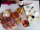 Platter of cured meats, cheeses, and drinks on a white surface with a bottle of wine.