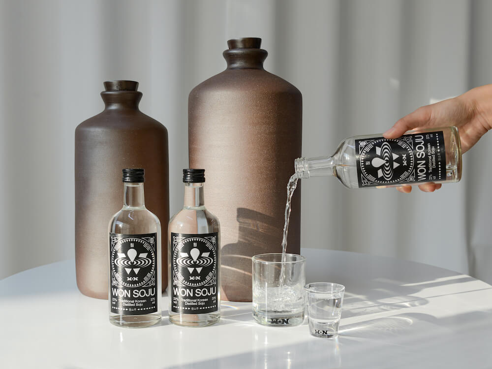Two bottles of Won Soju, a third being poured into a glass. Two large clay pots sit behind this tableau.