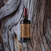 An appealing stately bottle of dark liquid with a burgandy foil wrapped top. The label is ornate in rich colors and reads: Del Professore Vermouth. There are large embossed letters of the same title running along the bottom of the bottle. This is set against the backdrop of a macro shot of a weathered looking tree, elegant lines and curves across the surface.