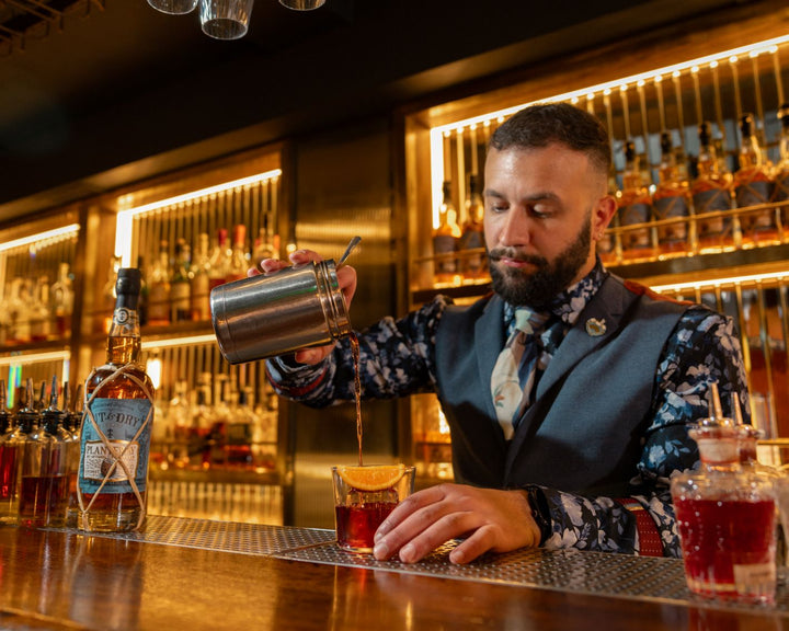 Bartender preparing a drink behind a bar with shelves of bottles in the background