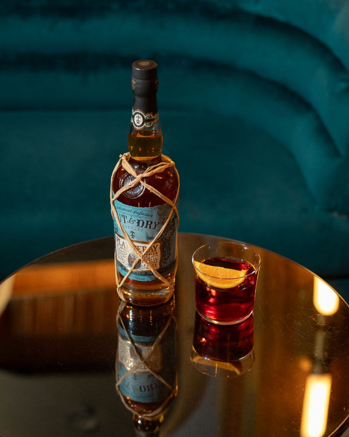 Bottle of Planteray Cut & Dry with a glass of a cocktail on a reflective surface against a teal background