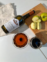 Calvados bottle, two glasses with a dark liquid and an orange slice, and a wooden cutting board with apples on a white surface.