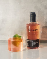 A bottle of Empirical Symphony 6 with a pink hued cocktail with a gree leaf garnish next to it. The background is minimalist, brushed metal.