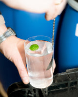 Close-up of a hand holding a glass with a clear liquid and a green leaf against a blue background.