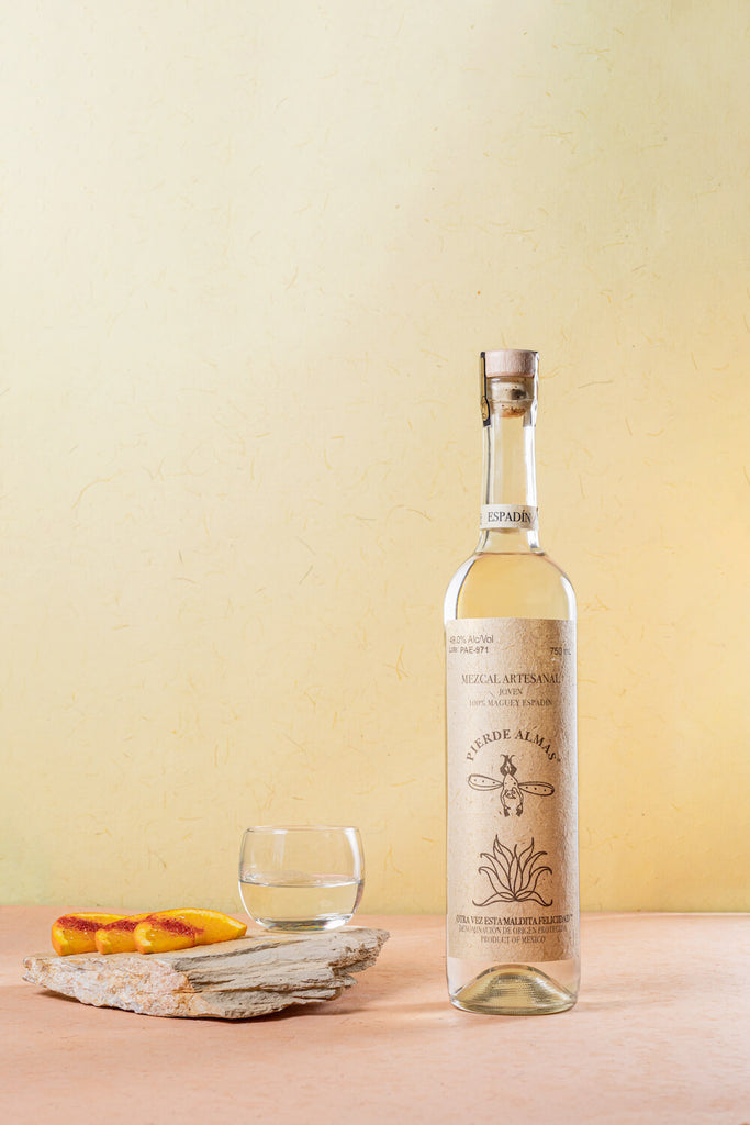 The tall bottle of Pierde Almas Mezcal to the left, a line of 3 orange slices dusted with taijin and a small glass filled with mezcal to the left.