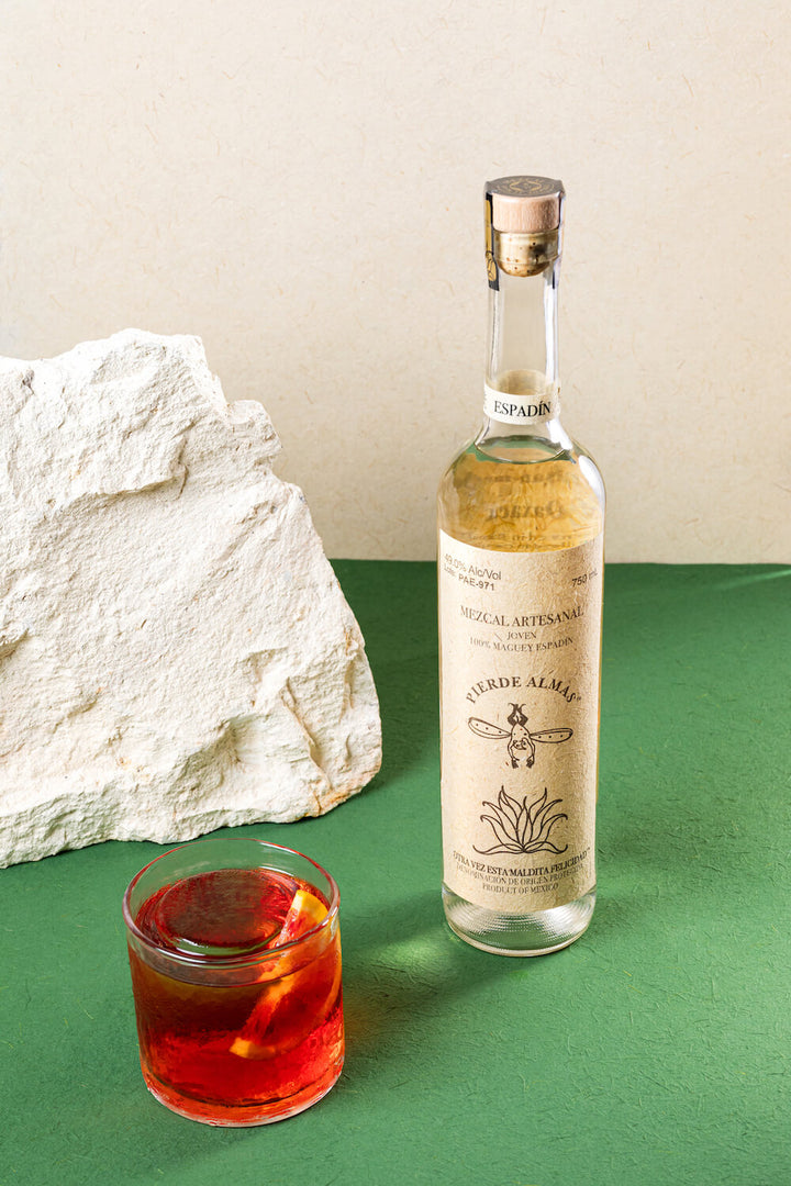 The tall bottle of Pierde Almas Mezcal to the right and a cocktail glass with a red liquid and a blood orange slice in it. All on a green and white backdrop.