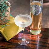 Cocktail glass and bottle of Orchard Pear liqueur on a wooden table with a plant and yellow napkin.