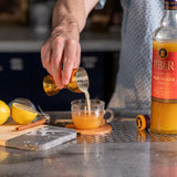 Person pouring a drink from a bottle labeled 'Liber' into a glass on a kitchen counter.
