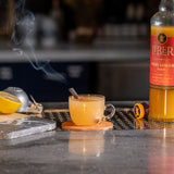 Hot beverage in a clear mug with steam, lemon wedge, and bottle of Liber & Co syrup on a bar counter.