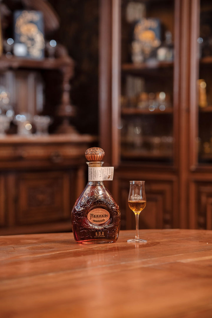 Bottle of Ferrand SDA Cognac with a glass of its contents on a wooden surface, with a bar setting in the background.